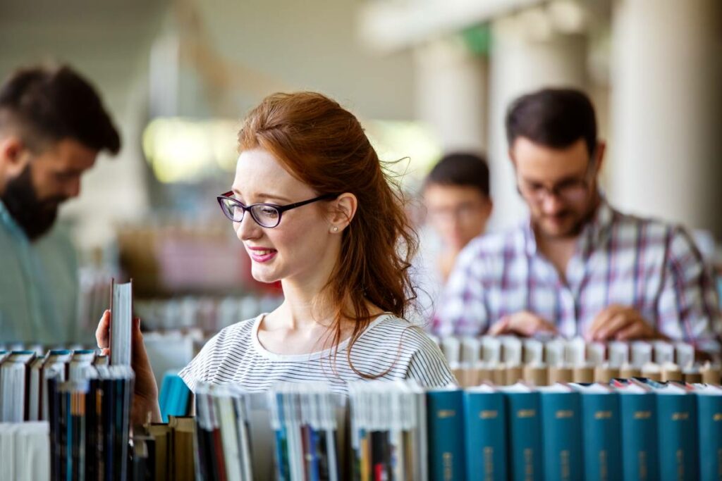 A line of books in the foreground with a few people in the background all looking down, presumably at books.