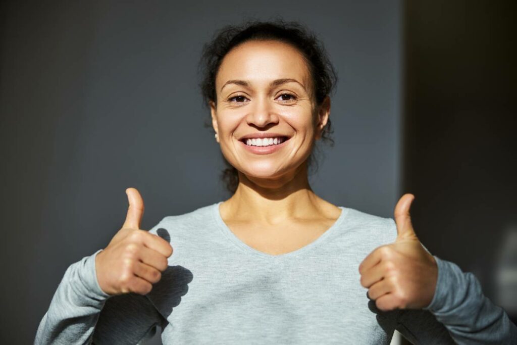 Latin-American female adult wearing a grey long sleeve shirt and giving two thumbs-up with a smile.